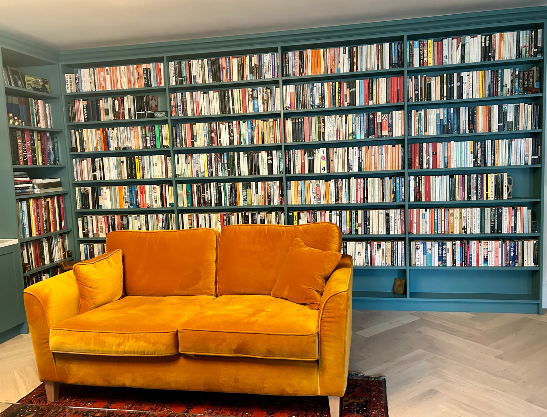 Alex Gunz’s home library, with teal bookshelves and a mustard sofa